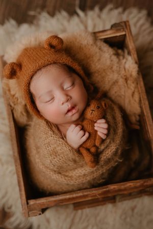 Sleeping newborn in cozy brown knit bear bonnet holding tiny teddy bear in wooden box.
