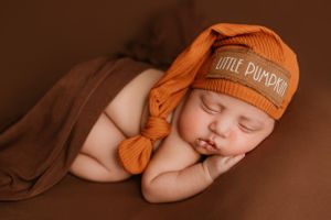 Peaceful newborn wearing orange "Little Pumpkin" beanie hat during autumn photoshoot.
