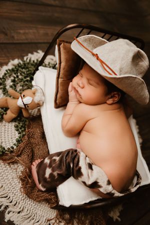 Sleeping newborn wearing cowboy hat with plush toys in rustic setting.