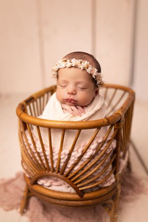 Sleeping newborn with flower crown wrapped in white swaddle inside vintage rattan chair.