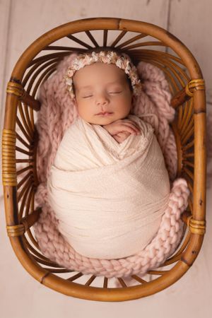 Newborn baby wrapped in white swaddling cloth sleeping peacefully in a rattan basket with pink knitted blanket.