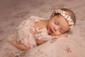 Peaceful sleeping infant wearing a lace outfit and floral crown on pink textured backdrop.