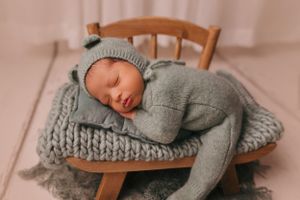 Small baby resting on wooden chair wearing grey knitted outfit and blanket.