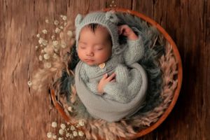 Sleeping newborn wrapped in soft gray blanket nestled in wooden bowl with baby's breath flowers.