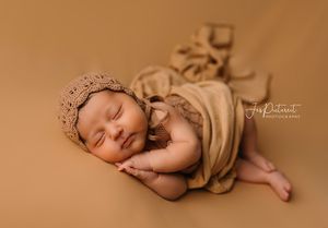 Sleeping baby in beige wrap and crown posed on warm brown background.