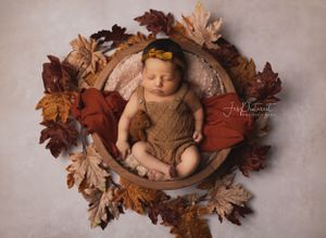 Sleeping baby in brown outfit surrounded by autumn leaves in wooden bowl.