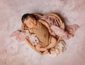 A sleeping infant in knit clothing resting in a heart-shaped basket with white flowers.