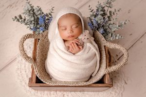 A sleeping newborn wrapped in white swaddle lies in a woven basket with blue flowers and eucalyptus decorations.