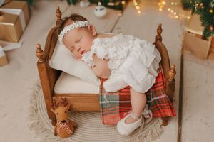 Sleeping baby in white dress on wooden doll bed with plaid blanket and Christmas decorations.