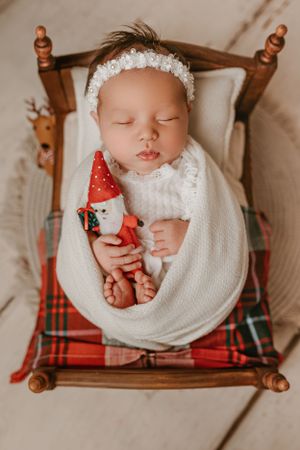 Sleeping infant wrapped in white with holiday decor on plaid blanket in wooden doll bed.