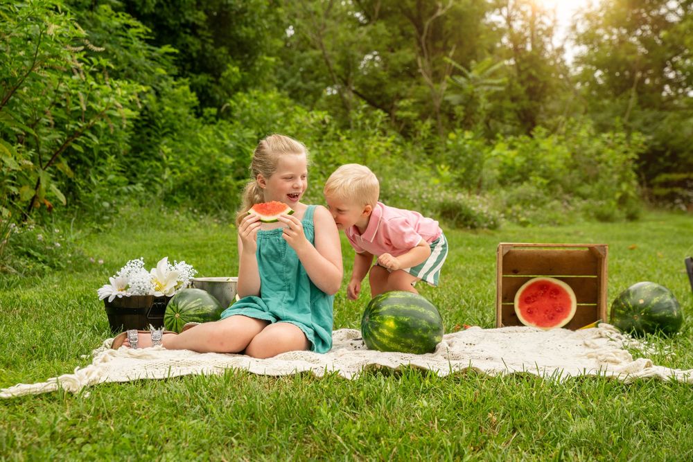 Two children enjoy watermelon slices at an outdoor picnic on green grass surrounded by whole watermelons and flowers.