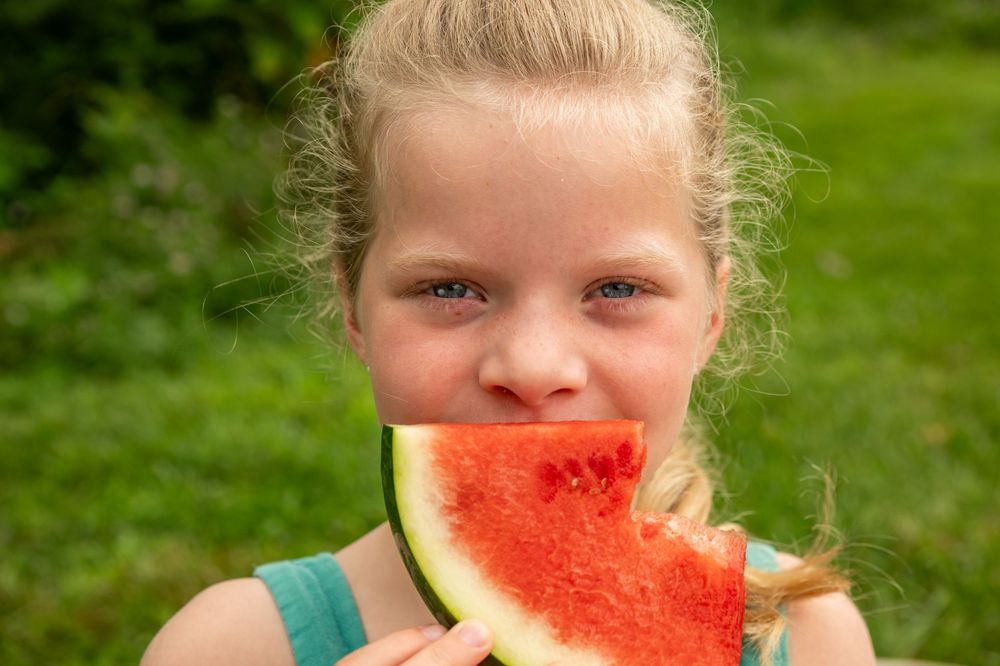 Blonde girl with blue eyes eating a fresh slice of red watermelon outdoors on a sunny summer day.