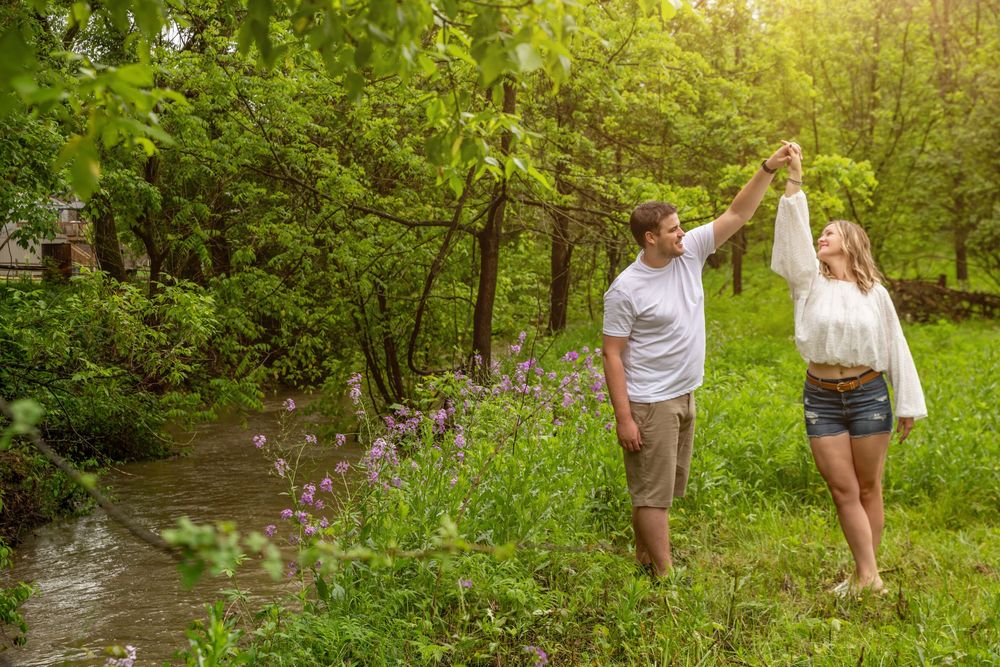Couple dancing outdoors near a creek surrounded by lush green trees and purple wildflowers in summer.
