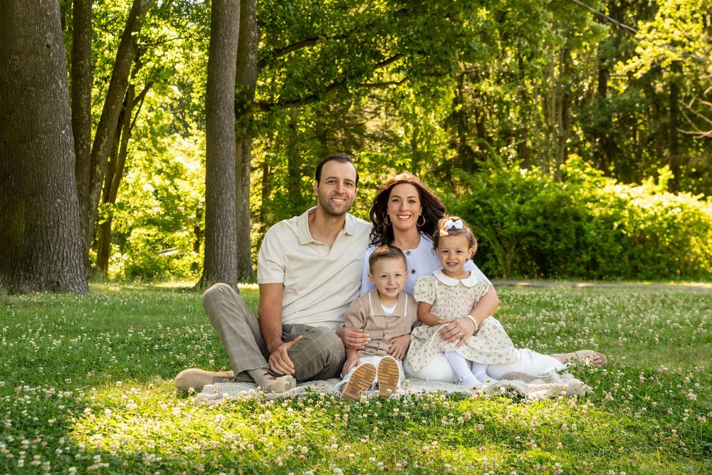 Family of four smiling on a blanket in a lush green park on a sunny day.