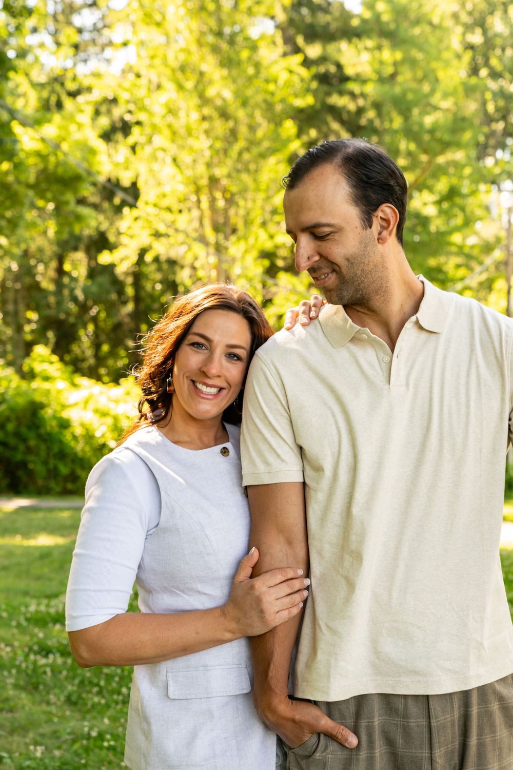 Happy couple embracing outdoors in a sunny green park, smiling woman in white dress with man in cream polo shirt.