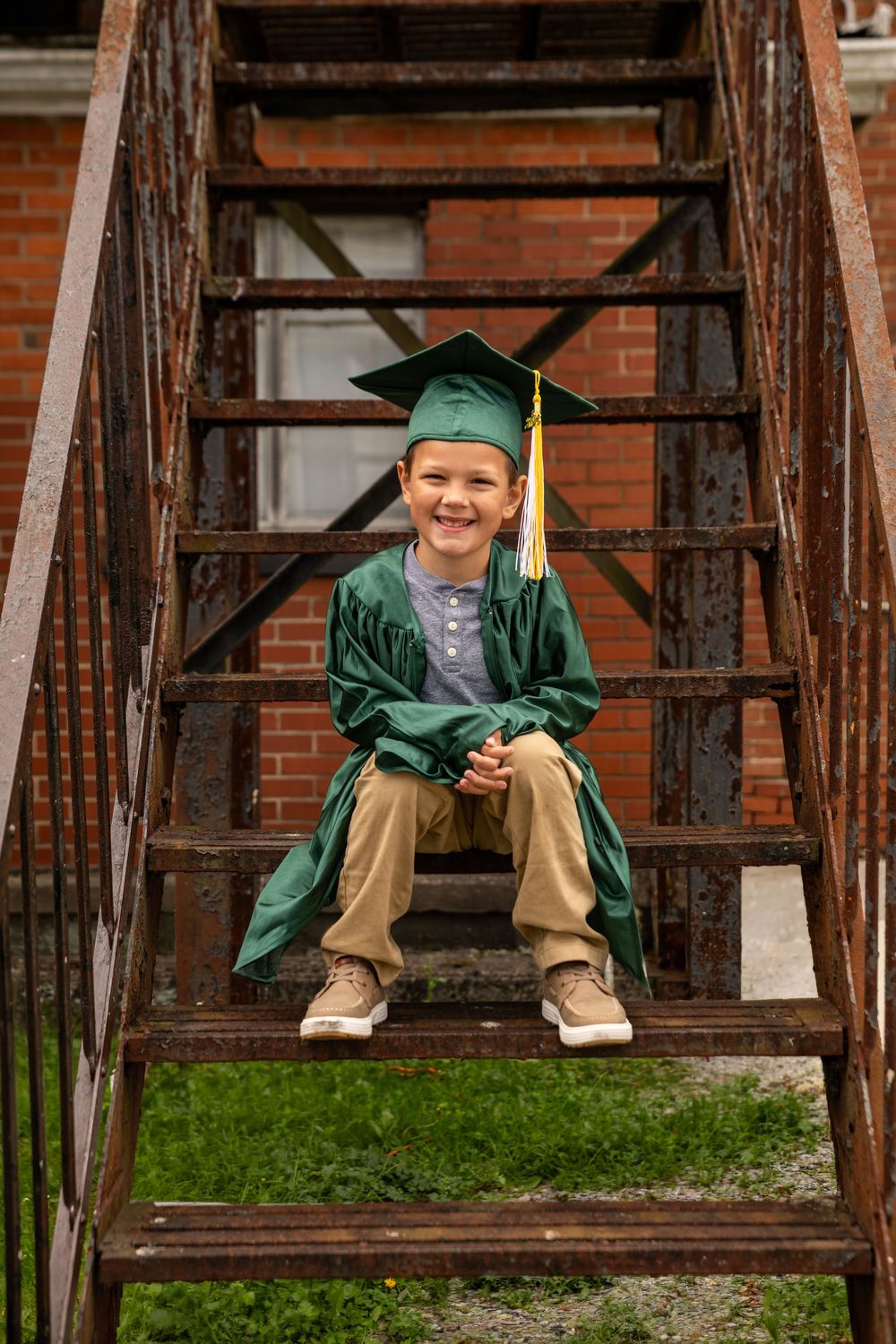 Smiling boy in green graduation cap and gown sitting on rustic metal stairs outdoors.