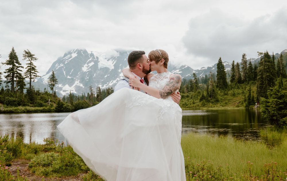 Panoramic mountain wedding scene with flowing white dress against evergreen forest and lake.