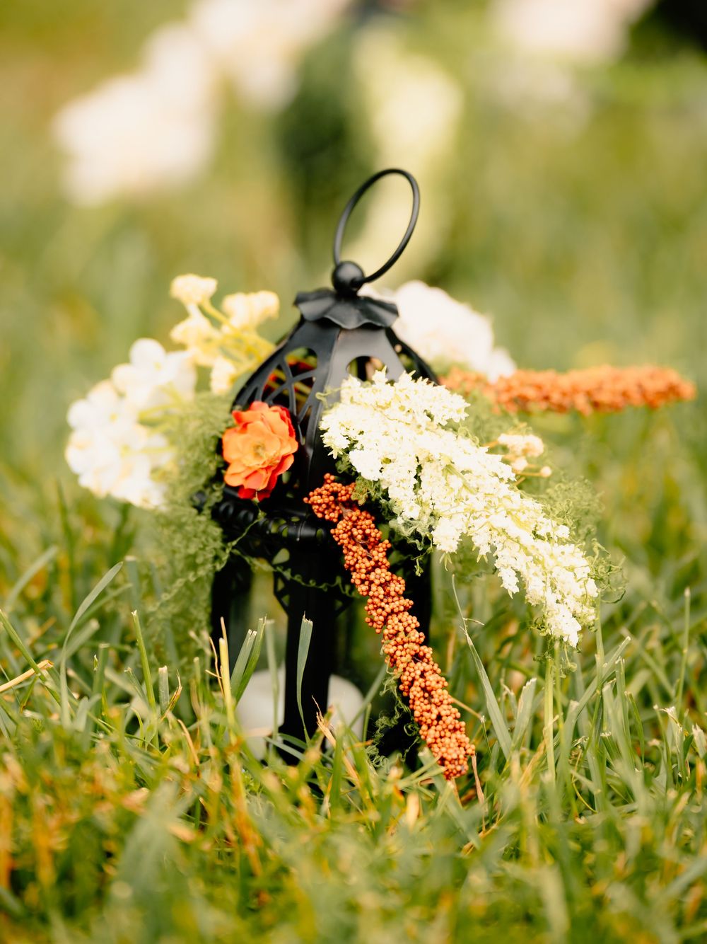 A black decorative lantern adorned with white and orange flowers sits in green grass.