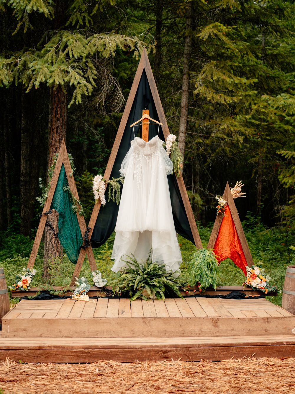 Multiple dresses displayed on wooden triangular arches on a wooden platform in an outdoor forest setting.