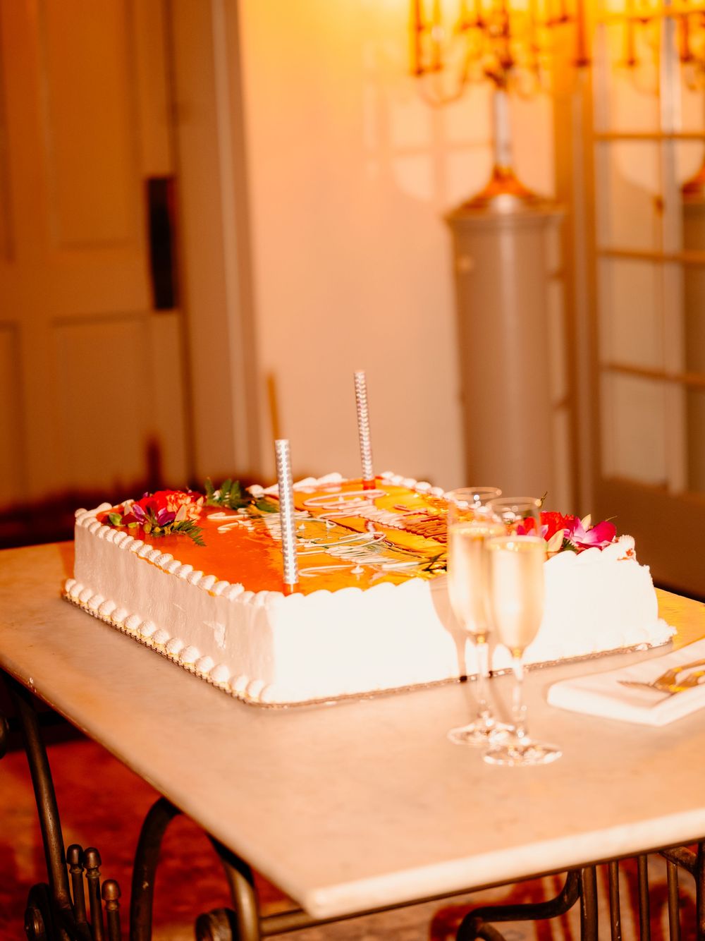 A rectangular wedding cake decorated with orange and pink flowers sits beside champagne glasses.