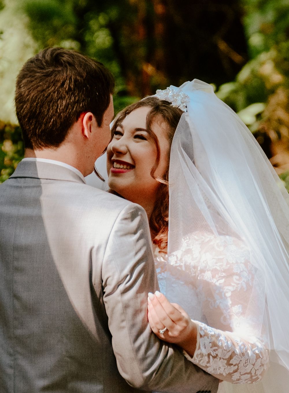 Wedding couple laughing together in sunlit garden wearing traditional white dress and gray suit.
