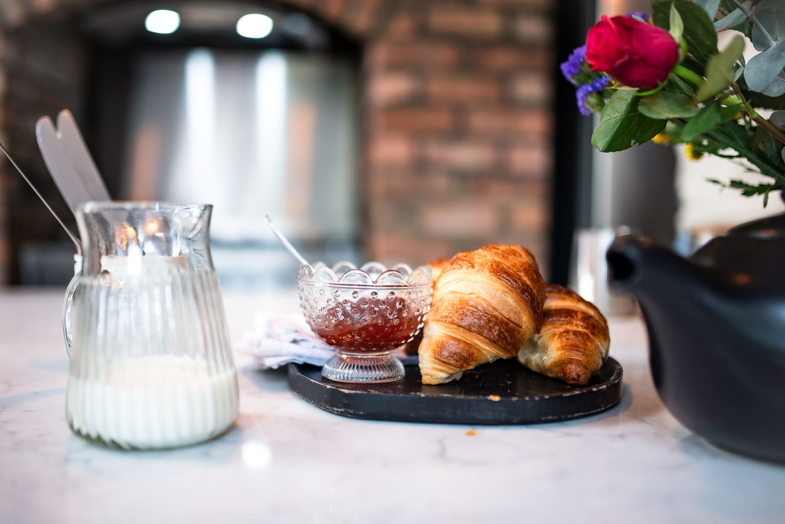 Fresh baked croissants served on dark slate with milk pitcher and red flowers on marble countertop in rustic setting.