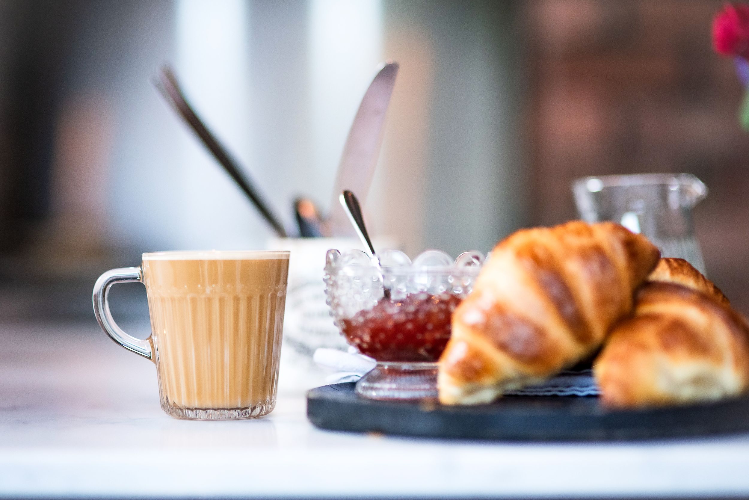 A coffee cup with steamed milk sits beside fresh croissants and jam on a dark plate against a blurred background.