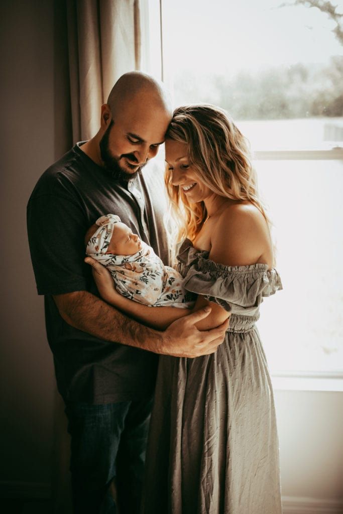 A couple in a sunlit room lovingly gazes at their newborn baby wrapped in a floral swaddle.