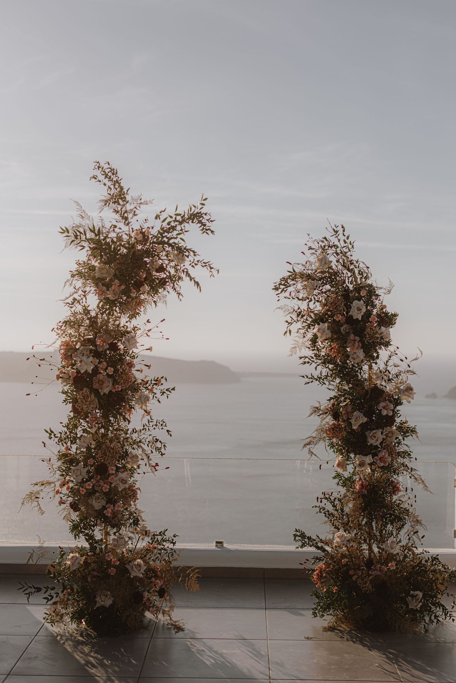 Tall floral arrangements frame a scenic ocean view at an outdoor wedding venue.