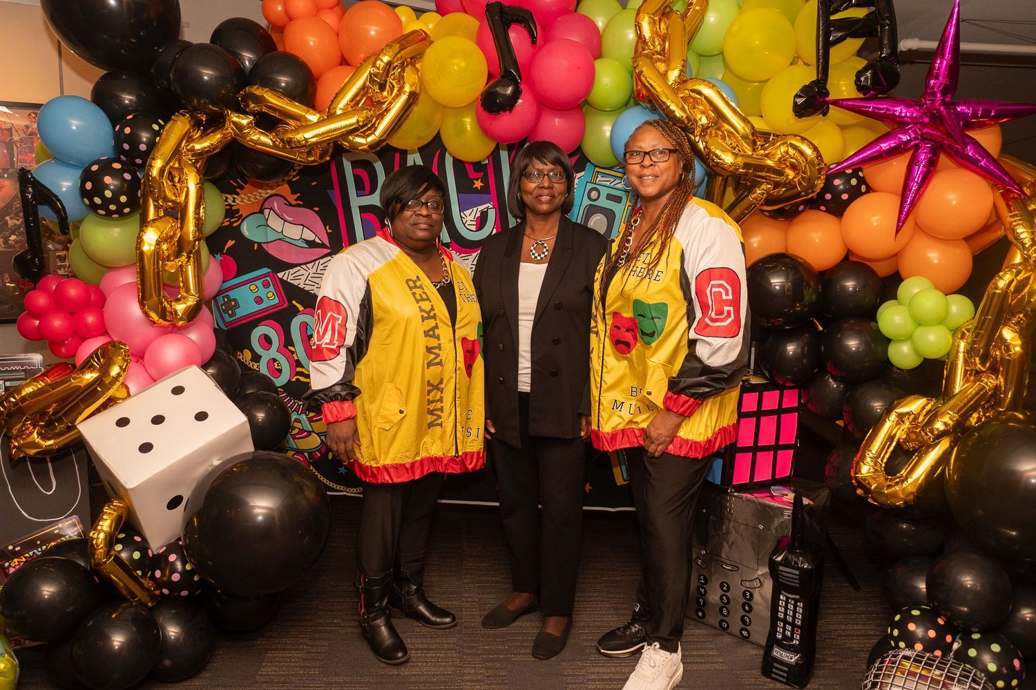 Three women pose in colorful varsity jackets at a festive party with balloon arch backdrop.