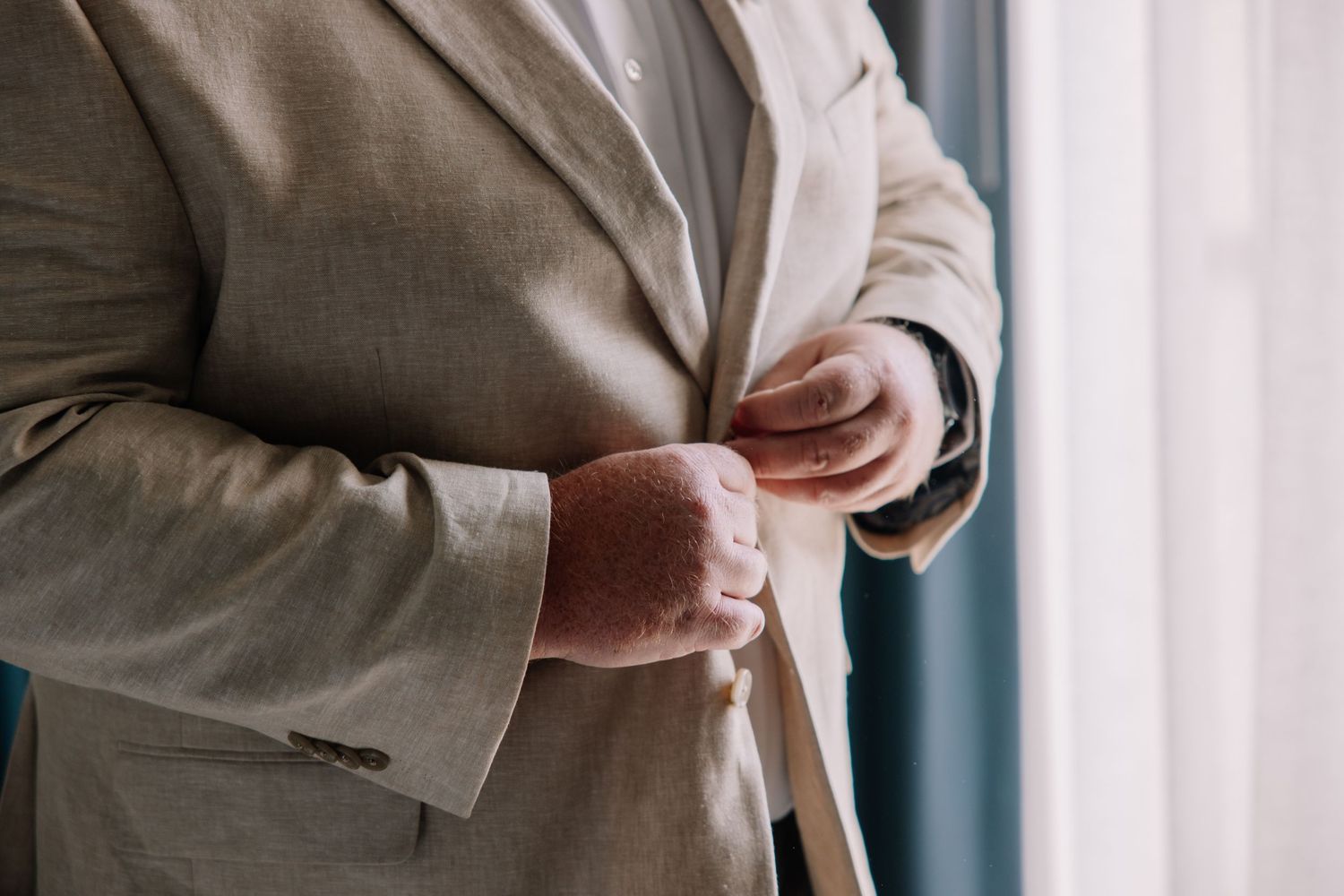 Close-up of a groom adjusting his tan suit jacket and boutonniere before a wedding ceremony.