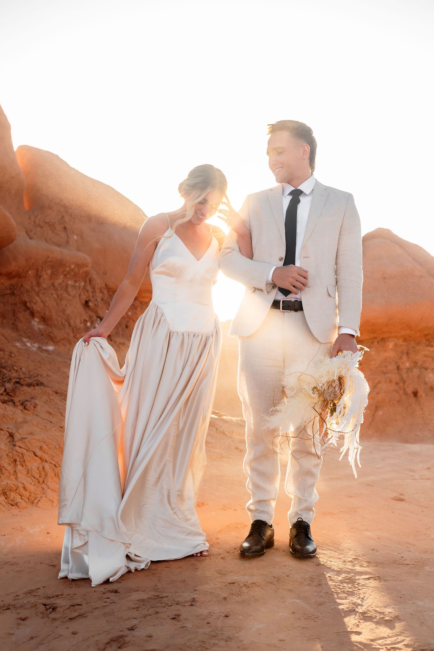 Wedding couple in white attire dances on desert rocks during sunset.