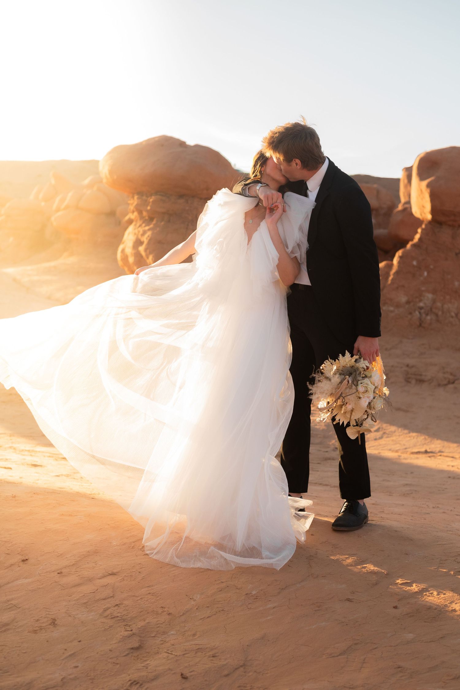 White wedding dress flows in desert breeze as couple poses among red rocks.