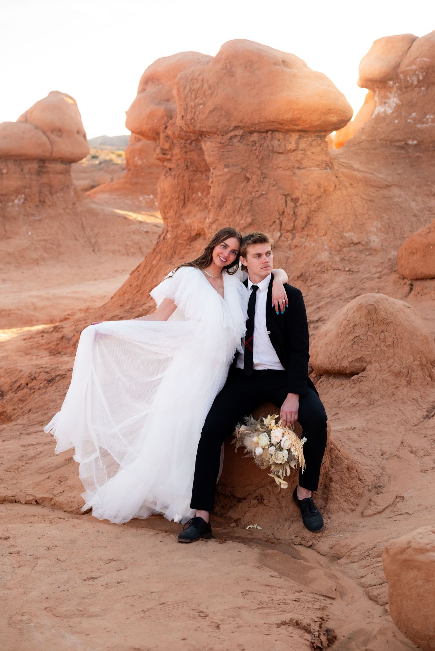 Wedding couple sits intimately against red rock formations in desert setting.