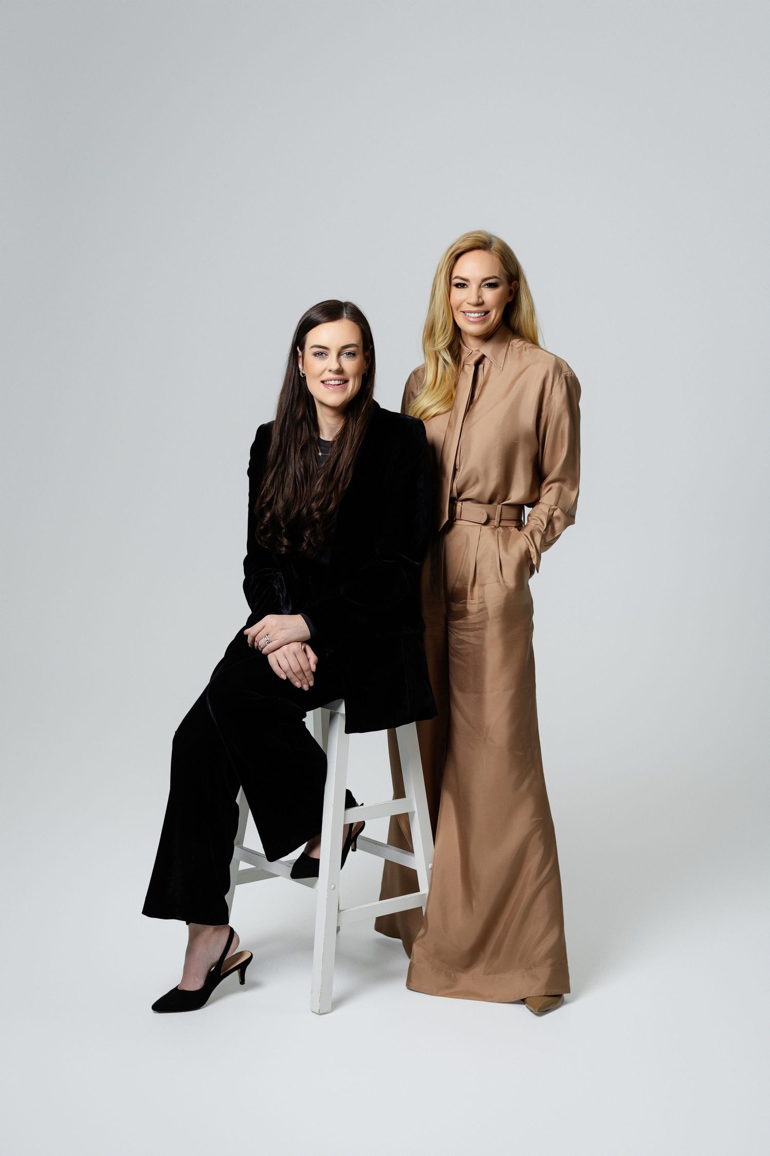 Two women pose in contrasting outfits - one in black and one in beige - against a light gray studio background.