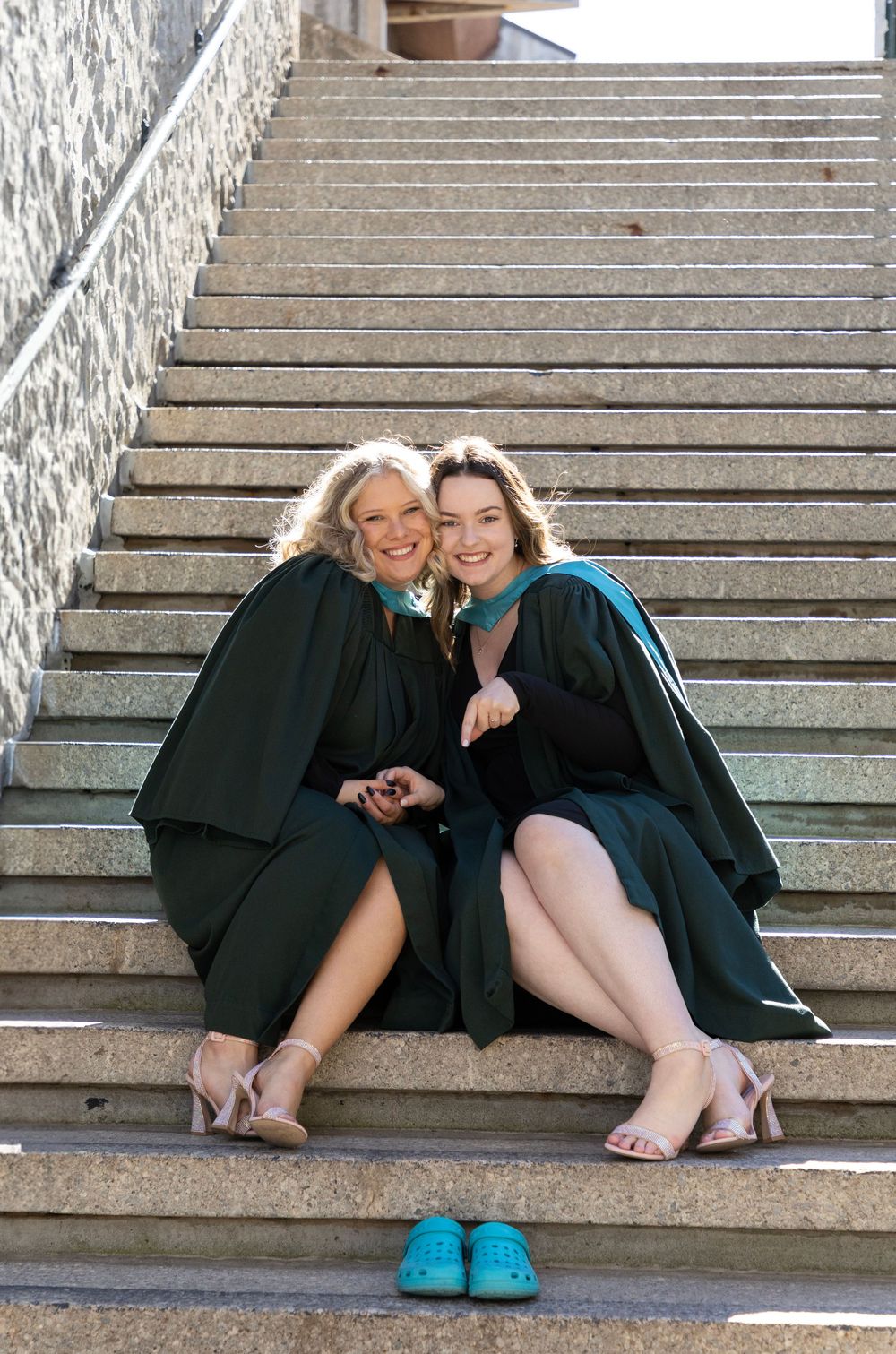 2 Female graduates of Trent University sitting on campus stairs wearing their gowns, pointing at a pair of blue crocs a couple steps below