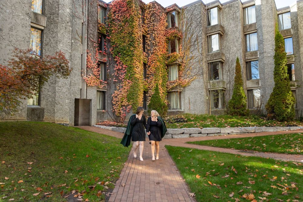 2 graduates walking and looking at each other at the trent champlain campus in the fall wearing their grad gowns draped over one shoulder