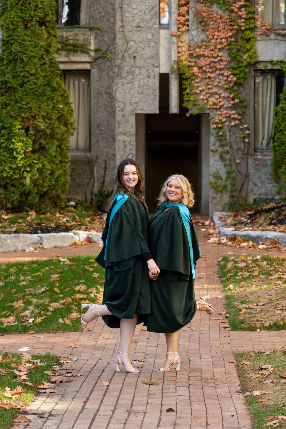 2 graduates standing next to each other at the trent champlain campus in the fall wearing their grad gowns with a leg popped out