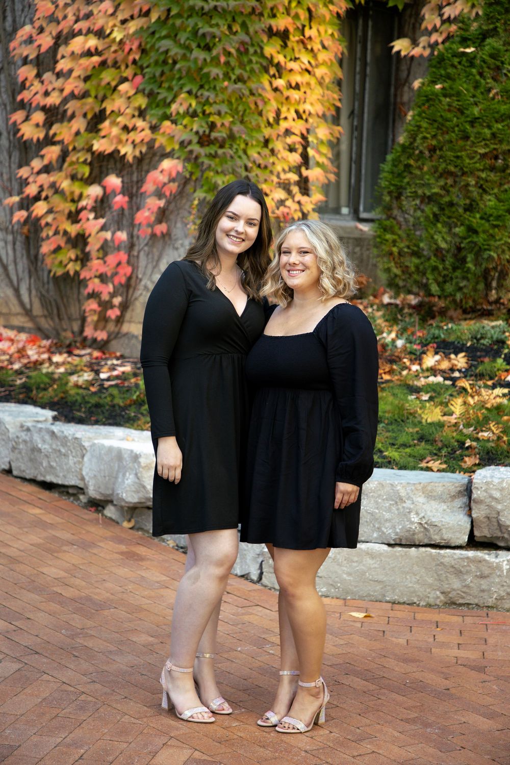 2 females wearing black dresses standing next to each other smiling at the camera (full body shot)