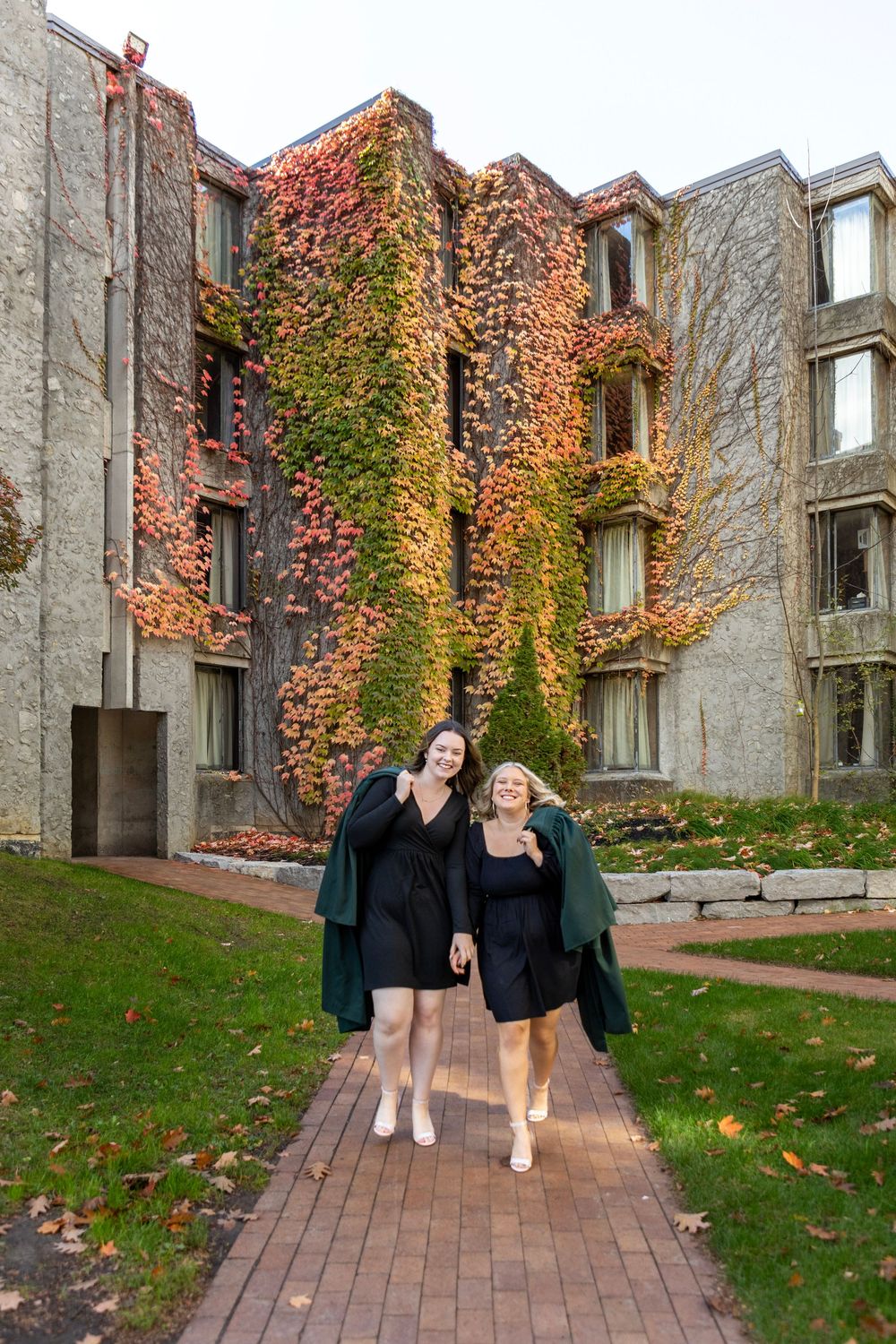 2 graduates walking and looking at each other at the trent champlain campus in the fall wearing their grad gowns draped over one shoulder