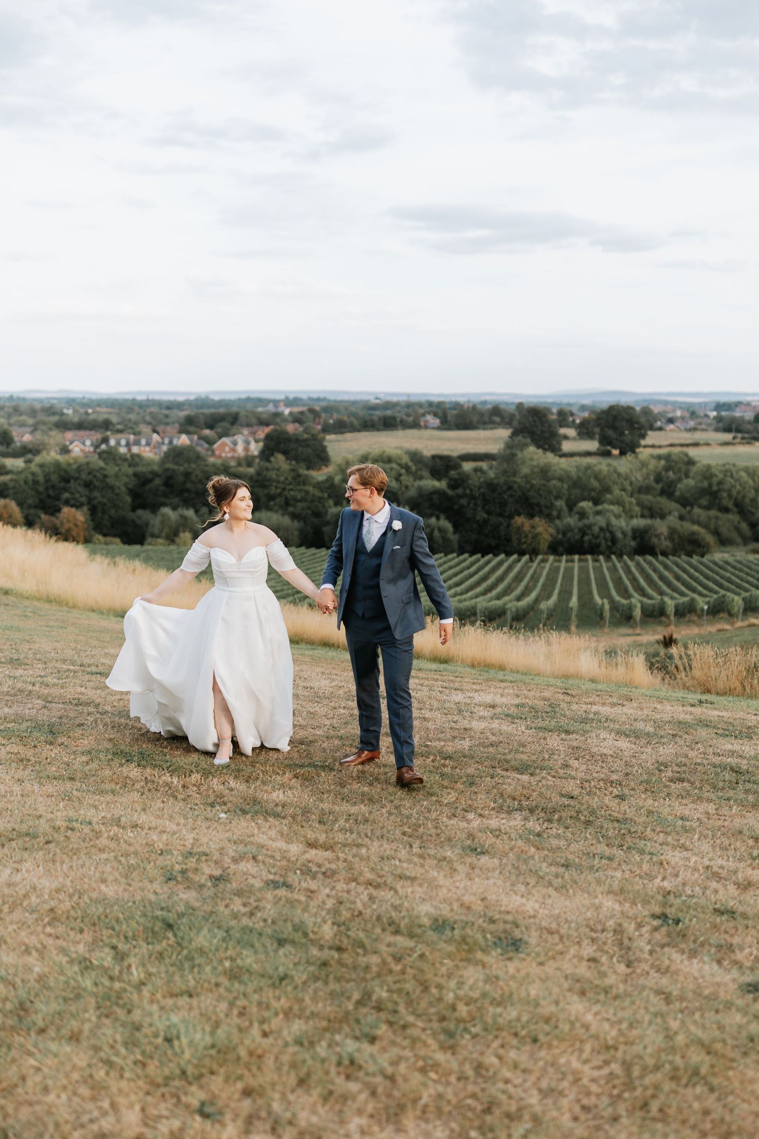 A couple walks through rolling countryside hills with scenic vineyard views in the background on their wedding day. Hencote Wedding