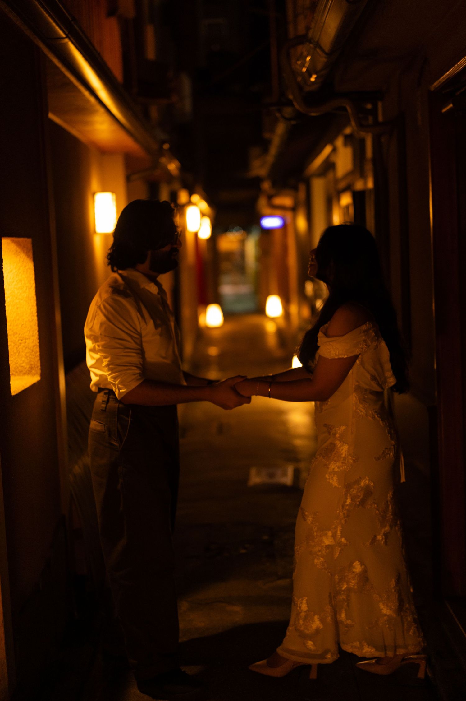 Couple holding hands in a glowing lantern-lit narrow alleyway at night in Japan.