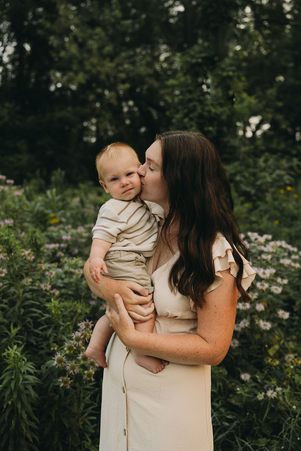 A tender moment captured outdoors as a mother in a white dress kisses her baby among blooming wildflowers and greenery.