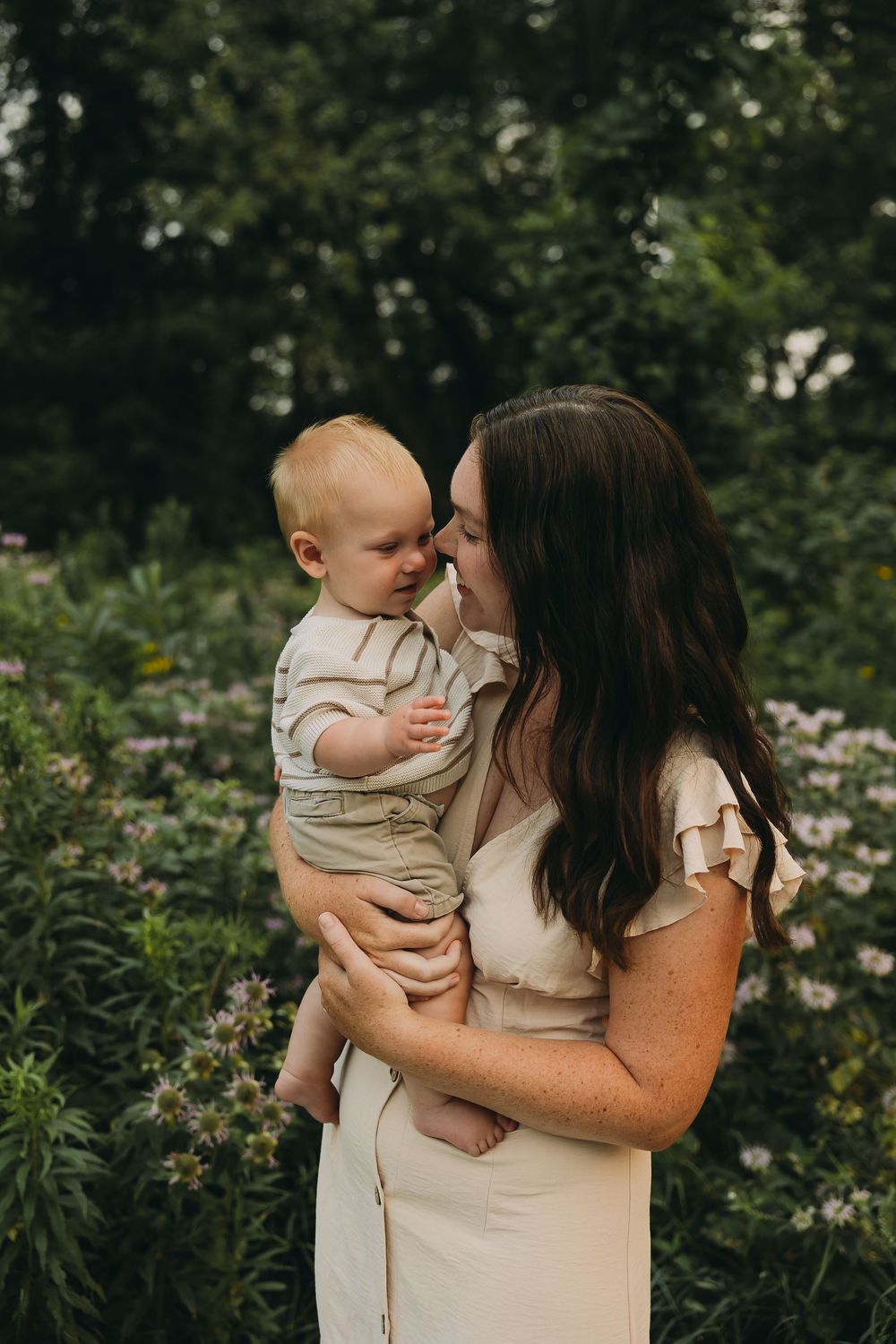 A tender moment between parent and baby outdoors among blooming flowers and greenery in a garden setting.