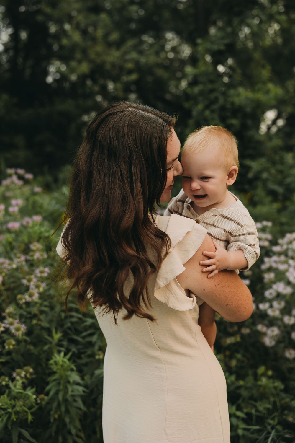 A tender moment captured in a garden setting as a parent in cream-colored clothing holds and embraces a small child.