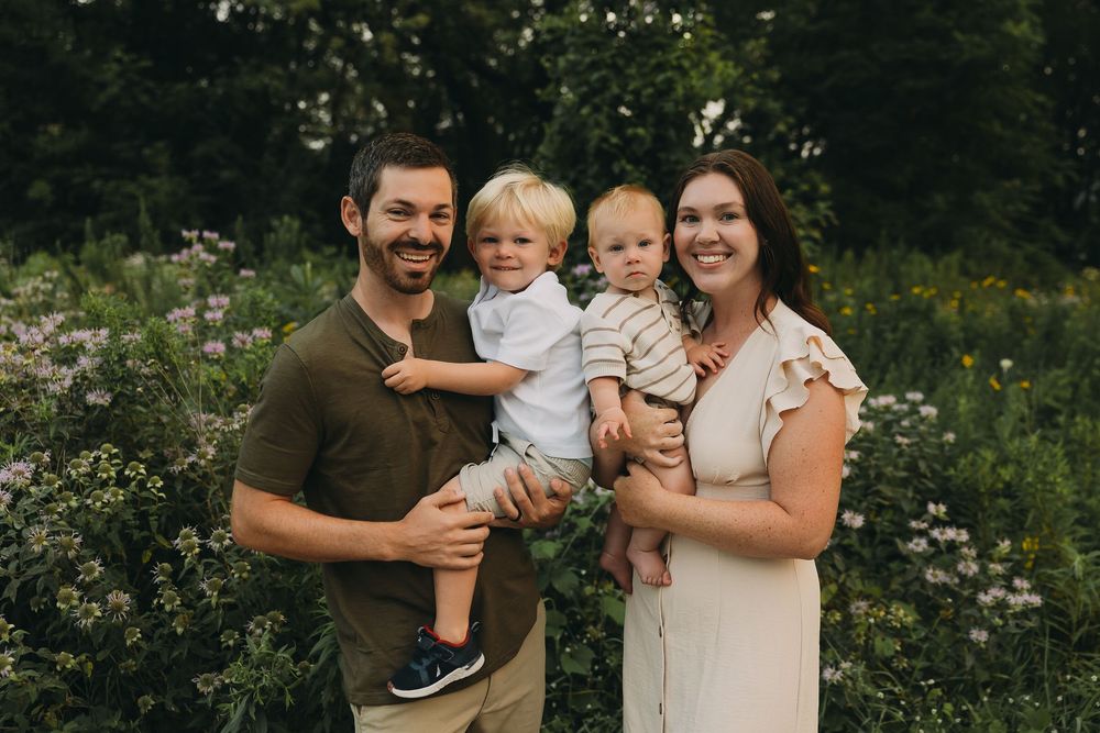 A family poses together outdoors in a meadow with white wildflowers on a sunny summer day.
