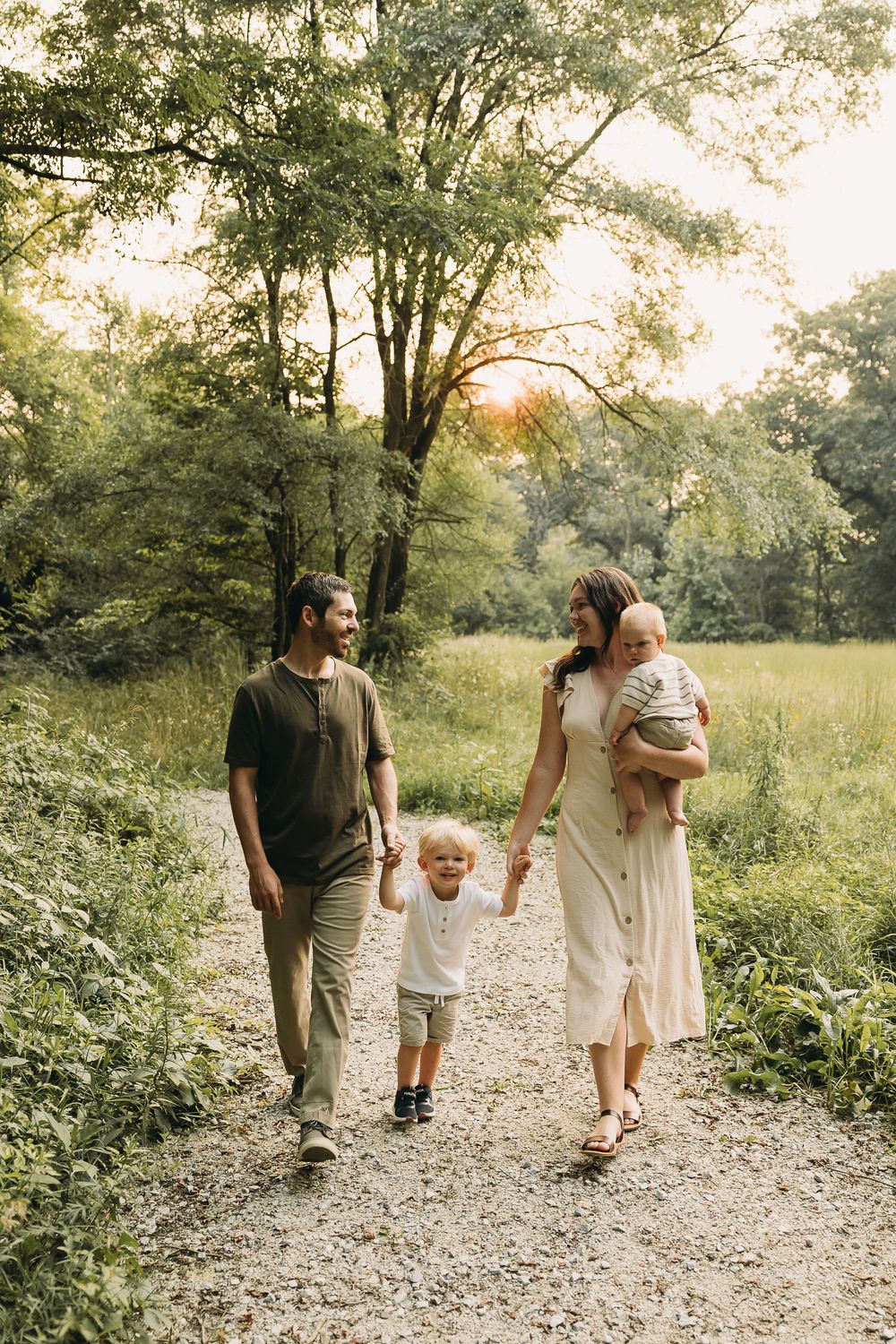 A family takes a stroll together on a gravel path through a sunlit wooded area on a warm afternoon.