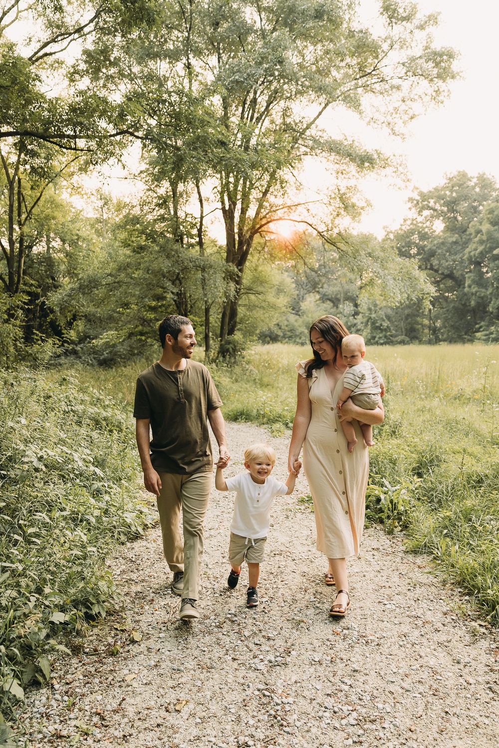 A family walks together on a sunlit gravel path through lush green woods during a summer evening.