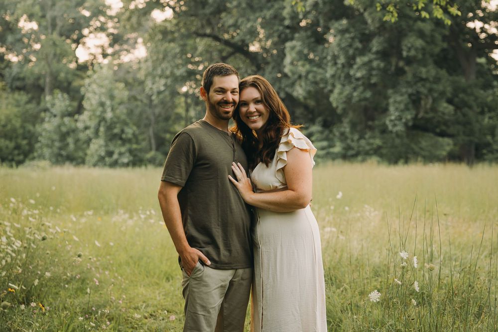 A couple embraces in a sunny meadow surrounded by tall grass and wildflowers.