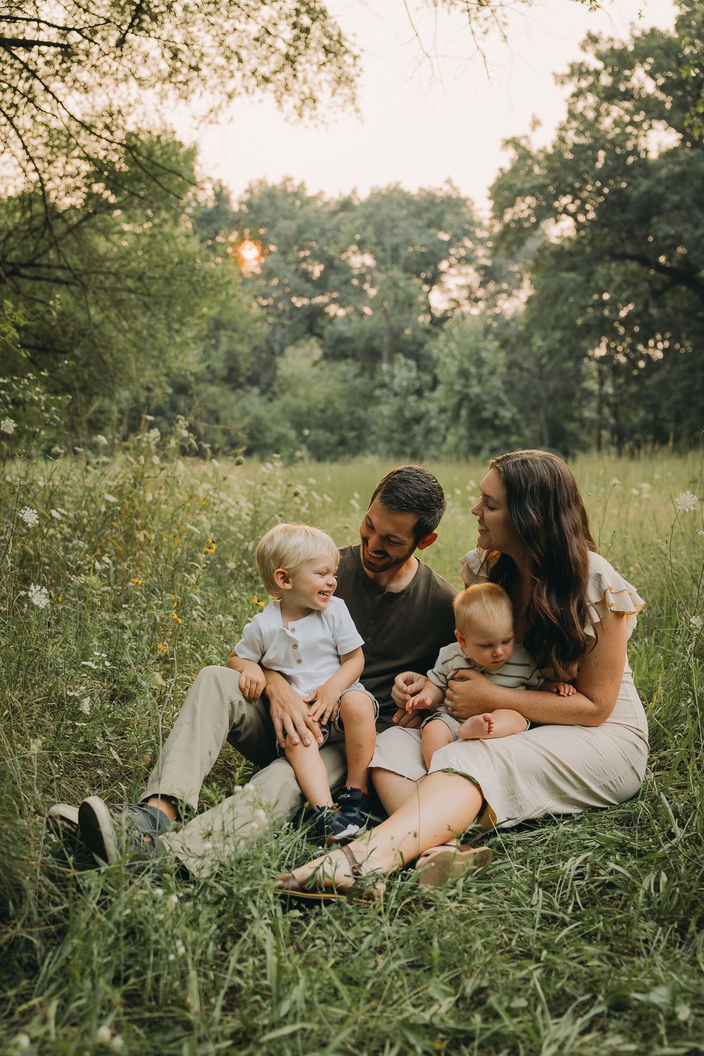 A family sits together in a sunlit meadow surrounded by tall grass and trees during golden hour.